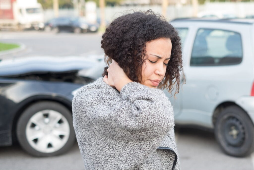 A woman holds her neck in pain while standing in front of two cars with visible collision damage in a parking lot.