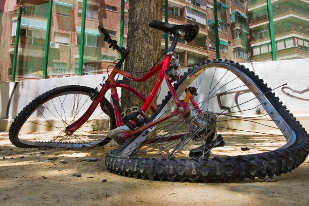 A damaged red bicycle with bent wheels lies on the ground under a tree near an apartment building and a green fence.