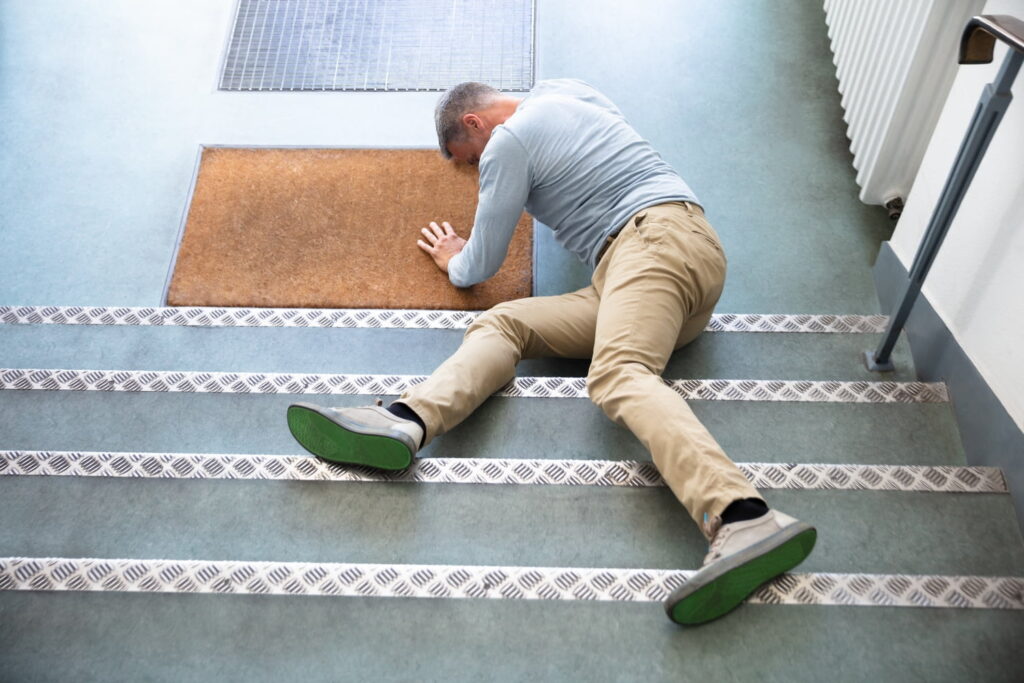 A man lies face down on a staircase, appearing to have fallen, with one arm supporting his upper body and legs positioned on different steps.
