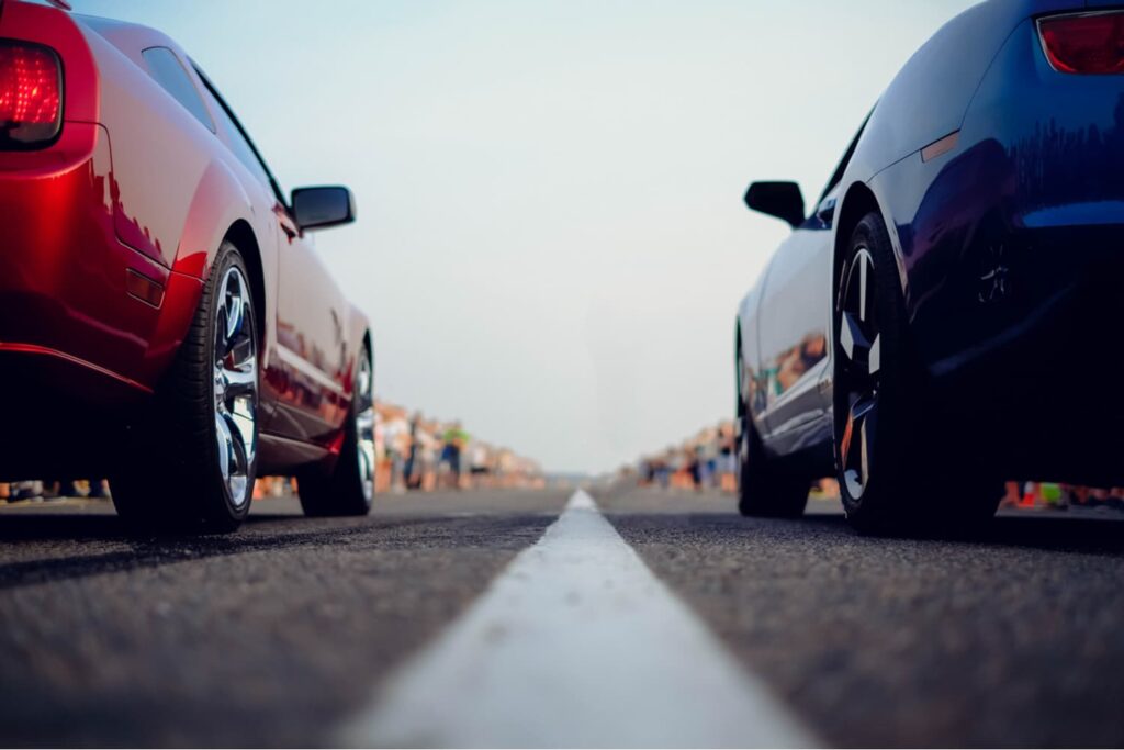 Two sports cars, one red and one blue, are positioned side by side on a road, viewed from behind and low to the ground, with a white line dividing the lanes.
