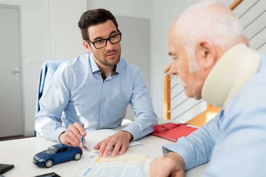 A man in a neck brace discusses paperwork with another man at a desk; a small model car and documents are visible on the table.