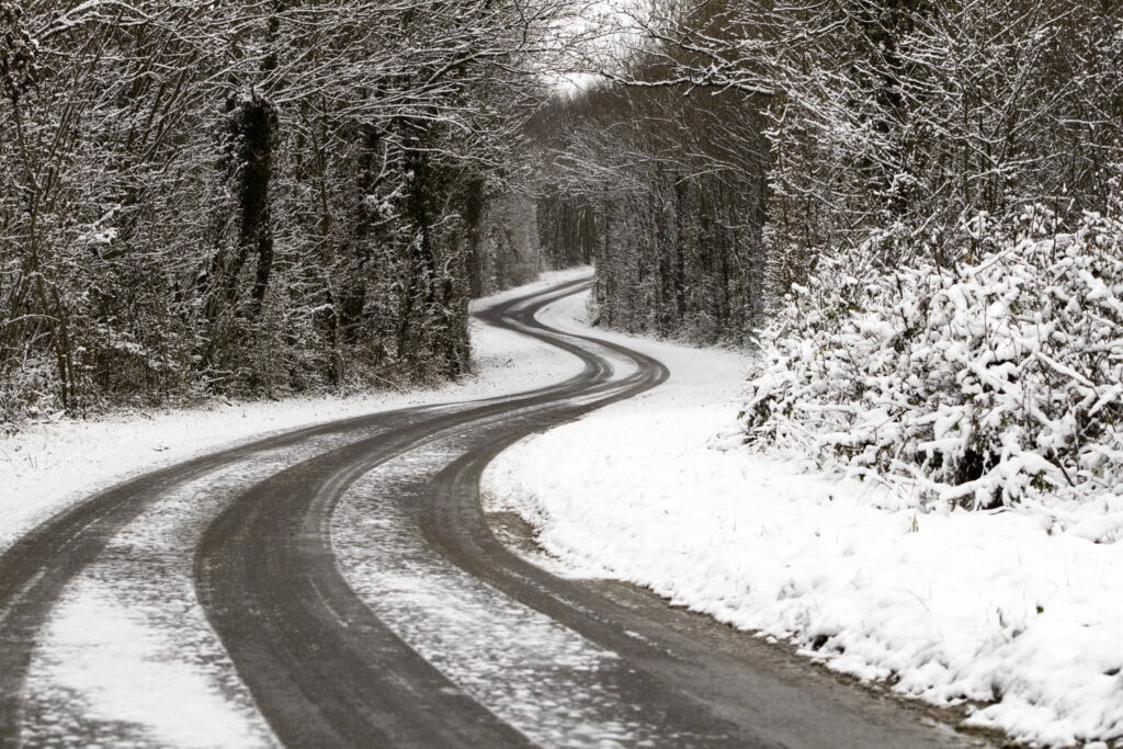 A winding road curves through a forest covered in snow, with tire tracks visible on the icy surface and bare trees lining both sides.