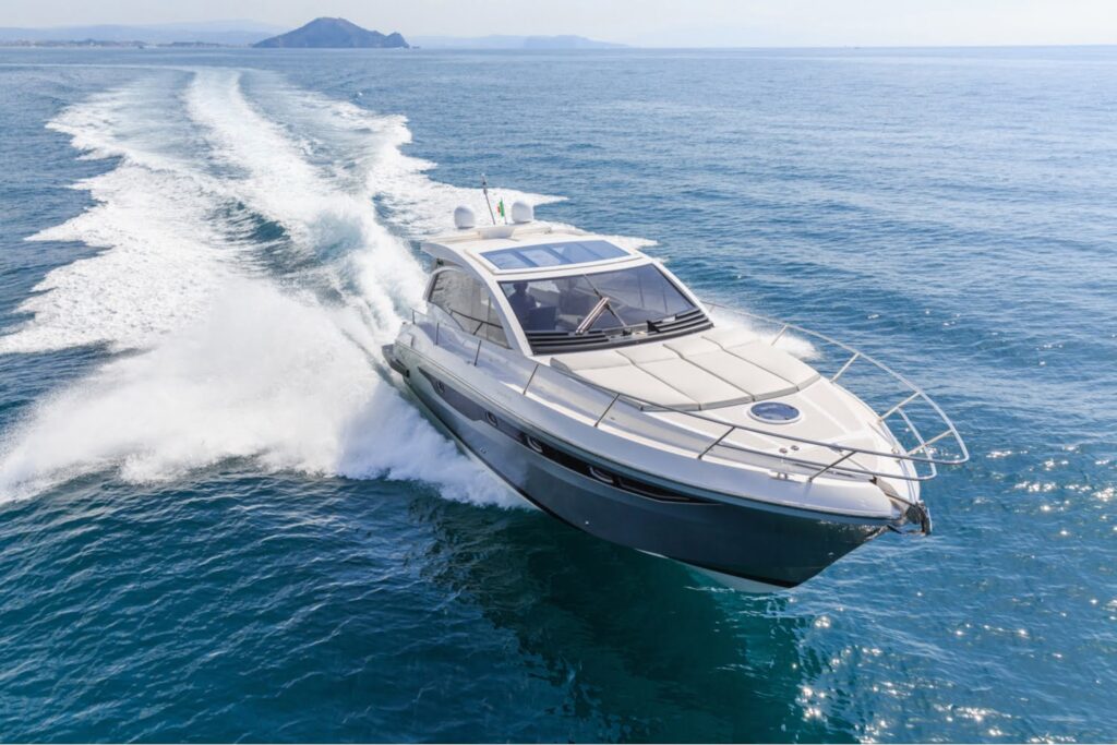 A modern motor yacht speeds across a calm, open sea, leaving a white wake behind, with distant land visible on the horizon.