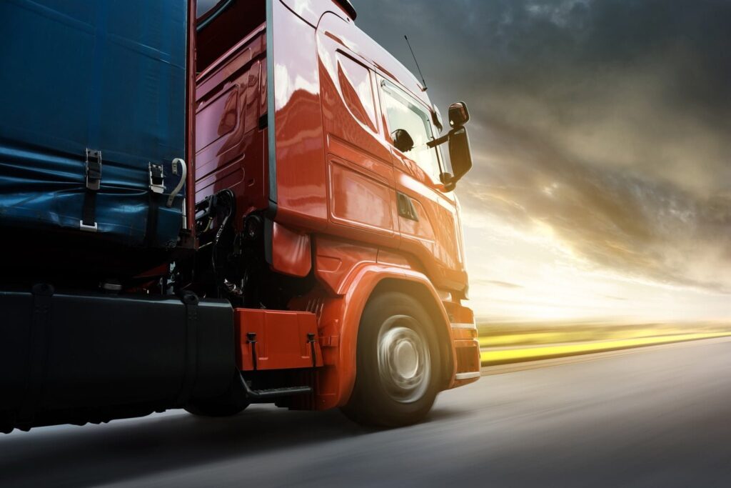 A red semi-truck with a blue trailer drives on a highway at sunset, with motion blur in the background indicating speed.