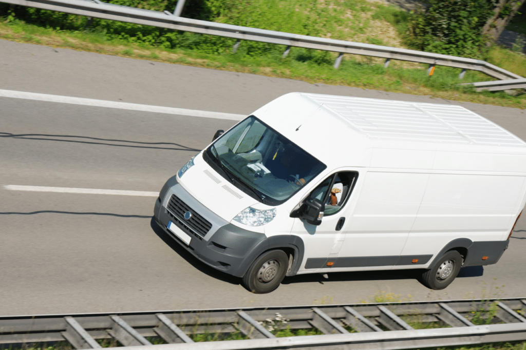 A white delivery van is driving on a multi-lane road, surrounded by guardrails and greenery.