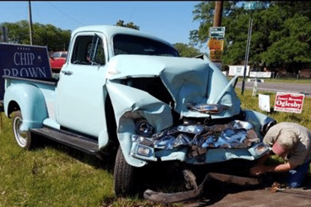 A light blue vintage truck with major front-end damage is parked on grass near campaign signs as a person kneels by the front tire.