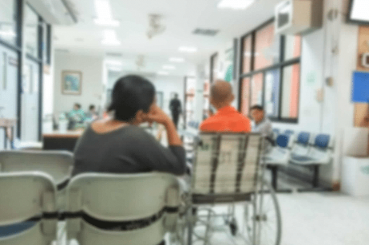Blurry image of people seated in a hospital waiting room, including one person in a wheelchair; medical staff and patients are visible in the background.