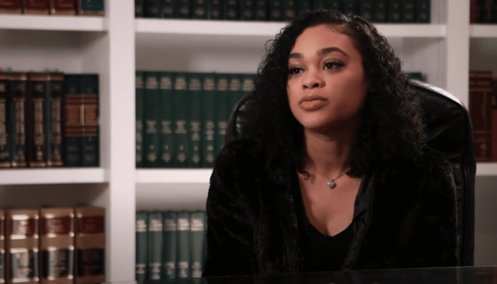 A woman with curly hair sits at a desk in front of bookshelves filled with legal books, looking slightly to the side.