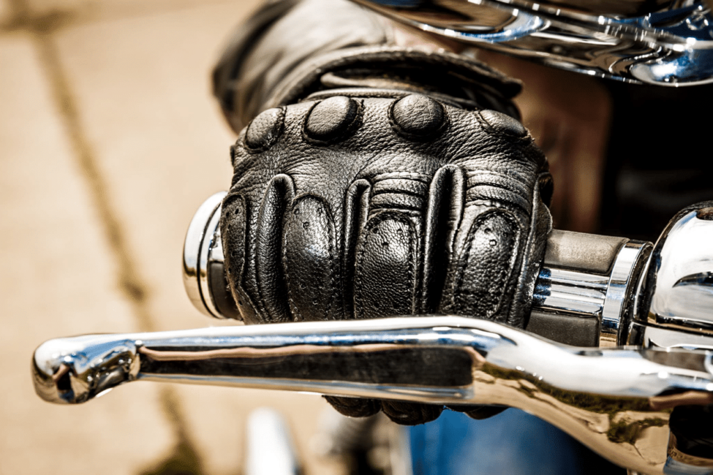 A close-up of a gloved hand gripping the handlebar of a motorcycle, with chrome details visible.