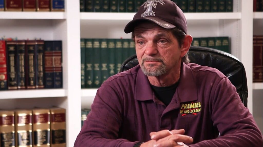 A man in a "Premier Driving Academy" shirt and cap sits at a desk with shelves of books in the background.