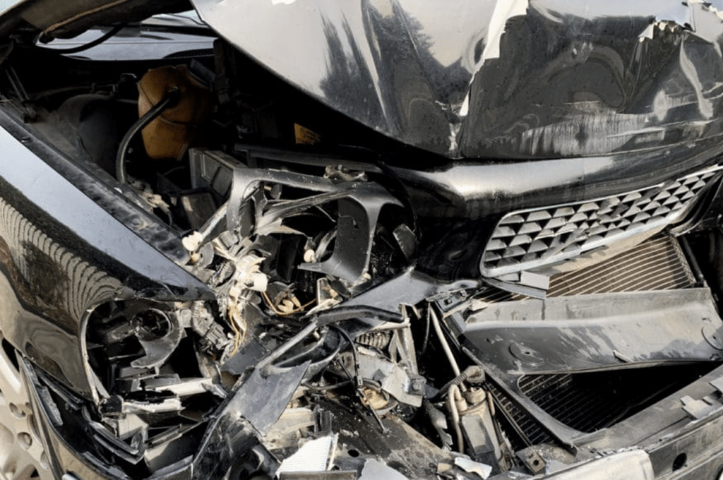 Close-up of the front end of a black car with severe damage from a collision, showing crumpled metal and exposed internal components.