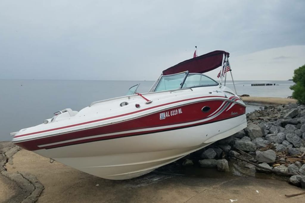 A white and red motorboat is beached on a rocky shoreline, partially out of the water, with a cloudy sky and calm sea in the background.
