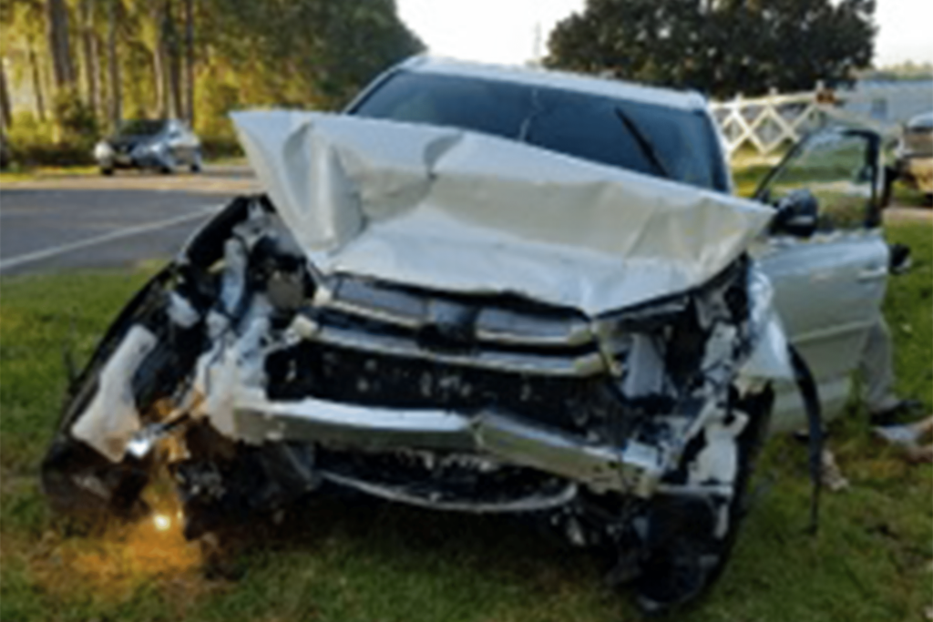 A car with severe front-end damage is parked on grass beside a road, with its hood crumpled and bumper detached.