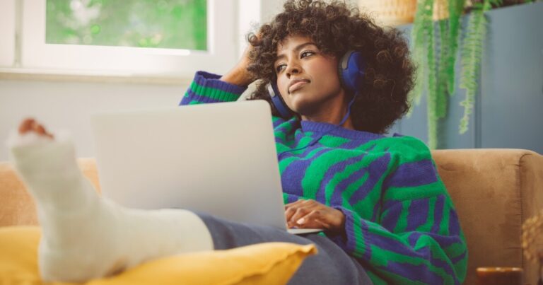 A person with headphones sits on a sofa with a bandaged foot elevated, using a laptop and looking thoughtfully to the side.