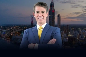 A man in a blue suit and yellow tie stands with arms crossed, smiling, in front of a cityscape at dusk.