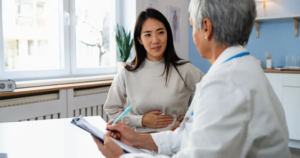 A woman holding her stomach speaks with a healthcare professional holding a clipboard in a medical office.