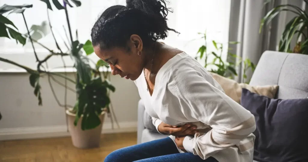 A woman sits on a couch, holding her stomach and leaning forward, appearing to be in discomfort or pain. There are several potted plants in the background.