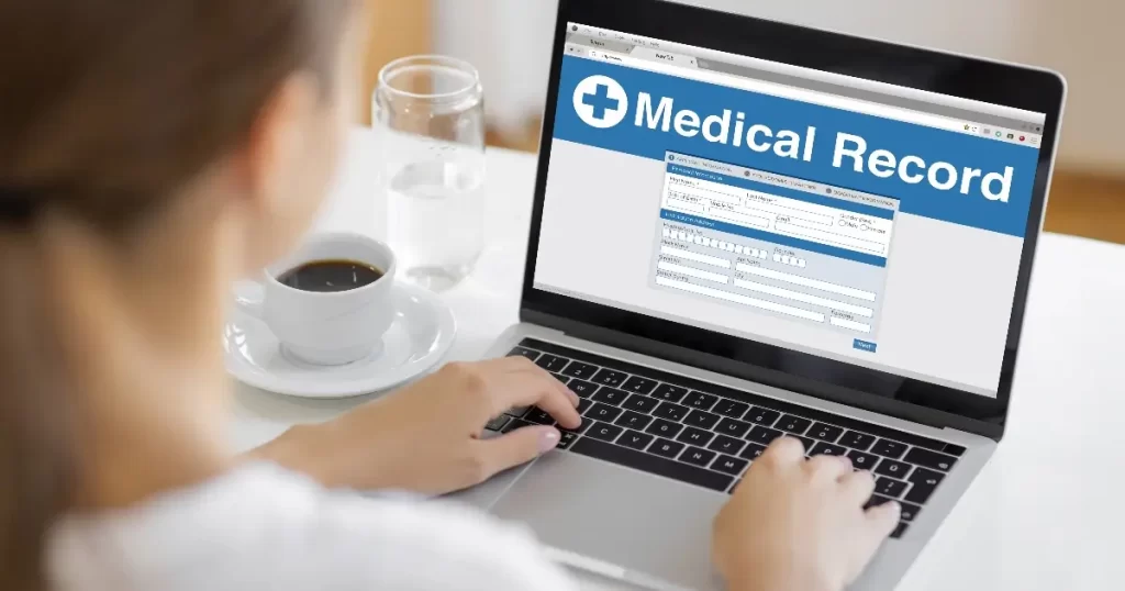 A person using a laptop displaying an electronic medical record form, with a coffee cup, glass of water, and notepad on the table.