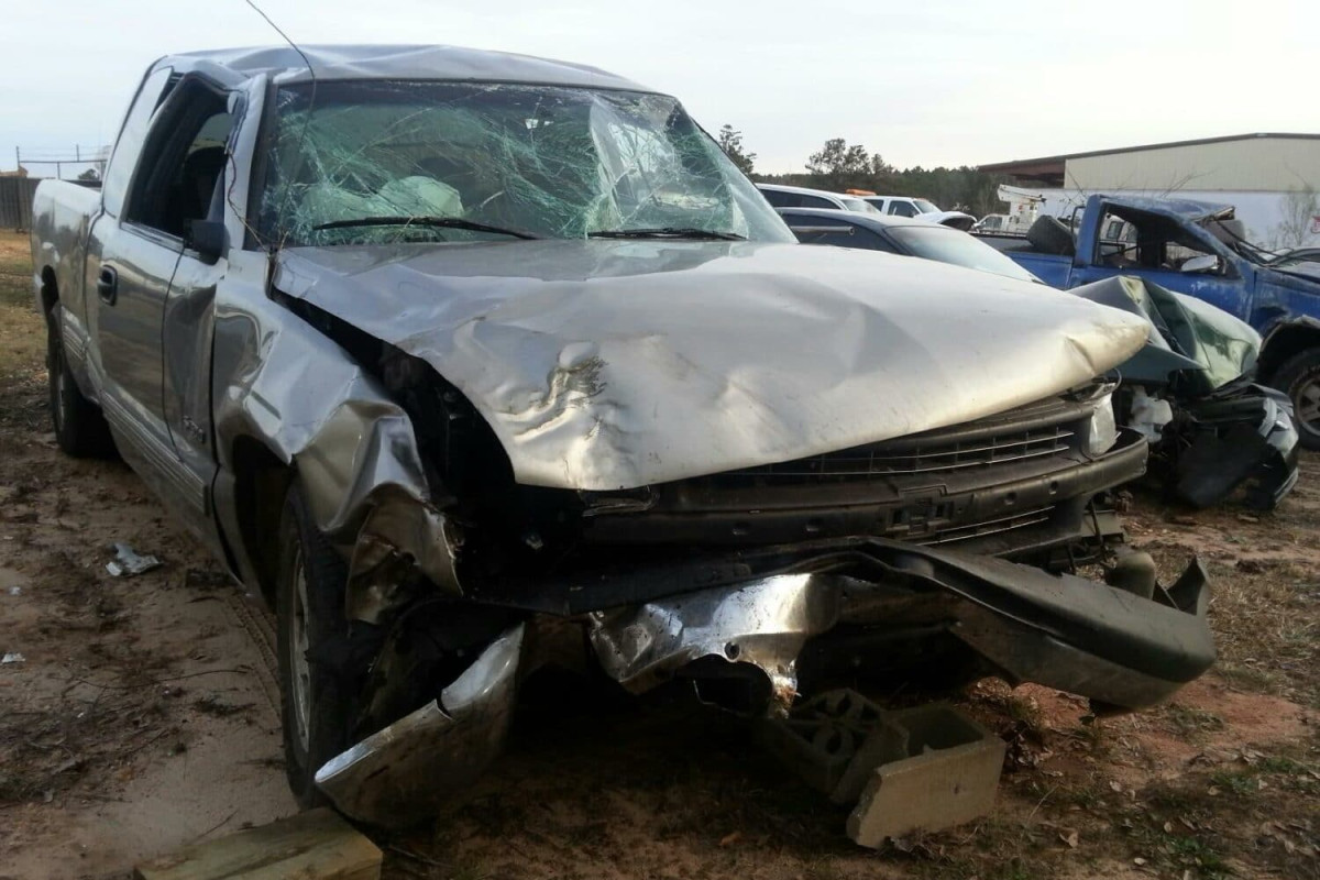 A heavily damaged silver pickup truck with a smashed front end and shattered windshield is parked outdoors near other vehicles.
