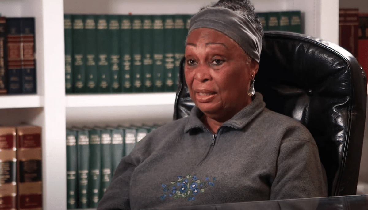 A woman wearing a gray headband and sweater sits in a black chair, speaking, with shelves of green and brown books in the background.
