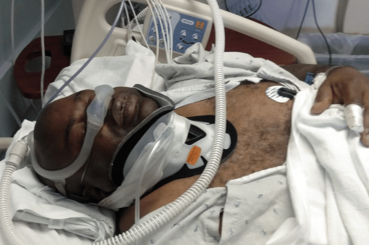 A person lies in a hospital bed with a neck brace, oxygen mask, and monitoring equipment attached to their chest and finger.