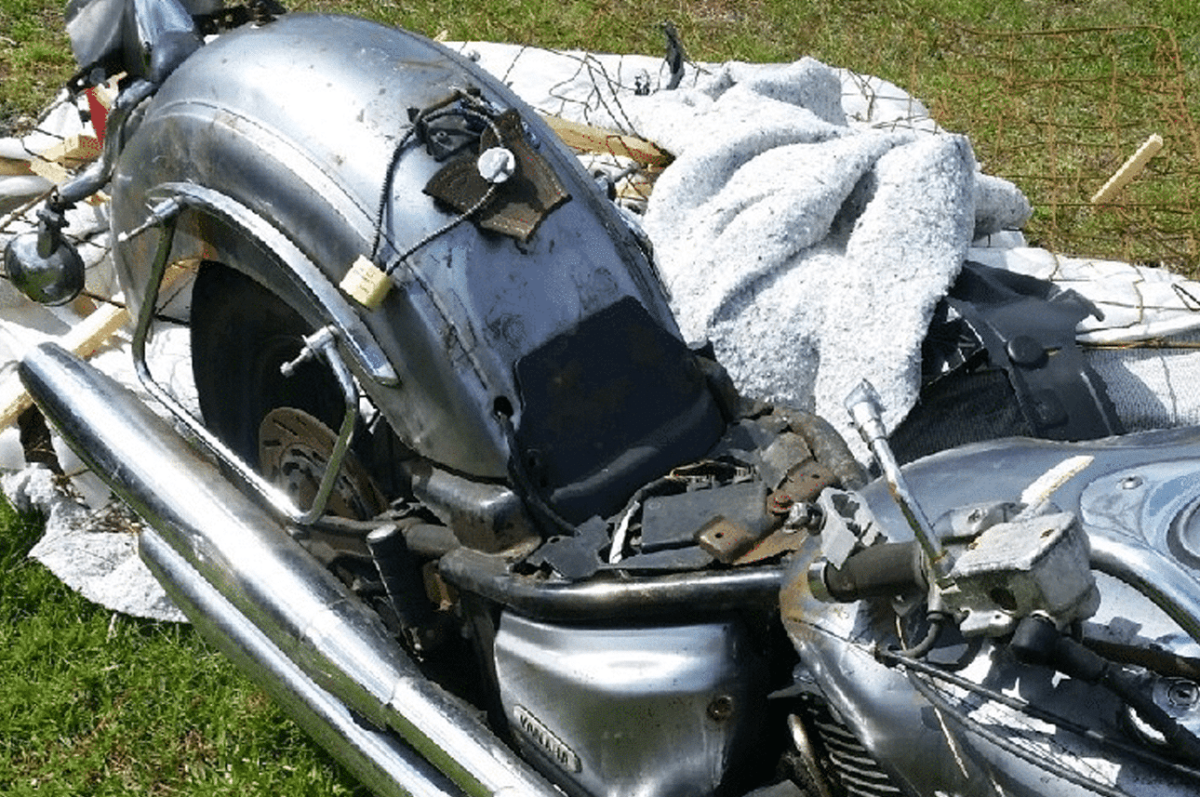A close-up of a damaged motorcycle lying on its side on grass, with parts missing or broken and debris scattered around it.