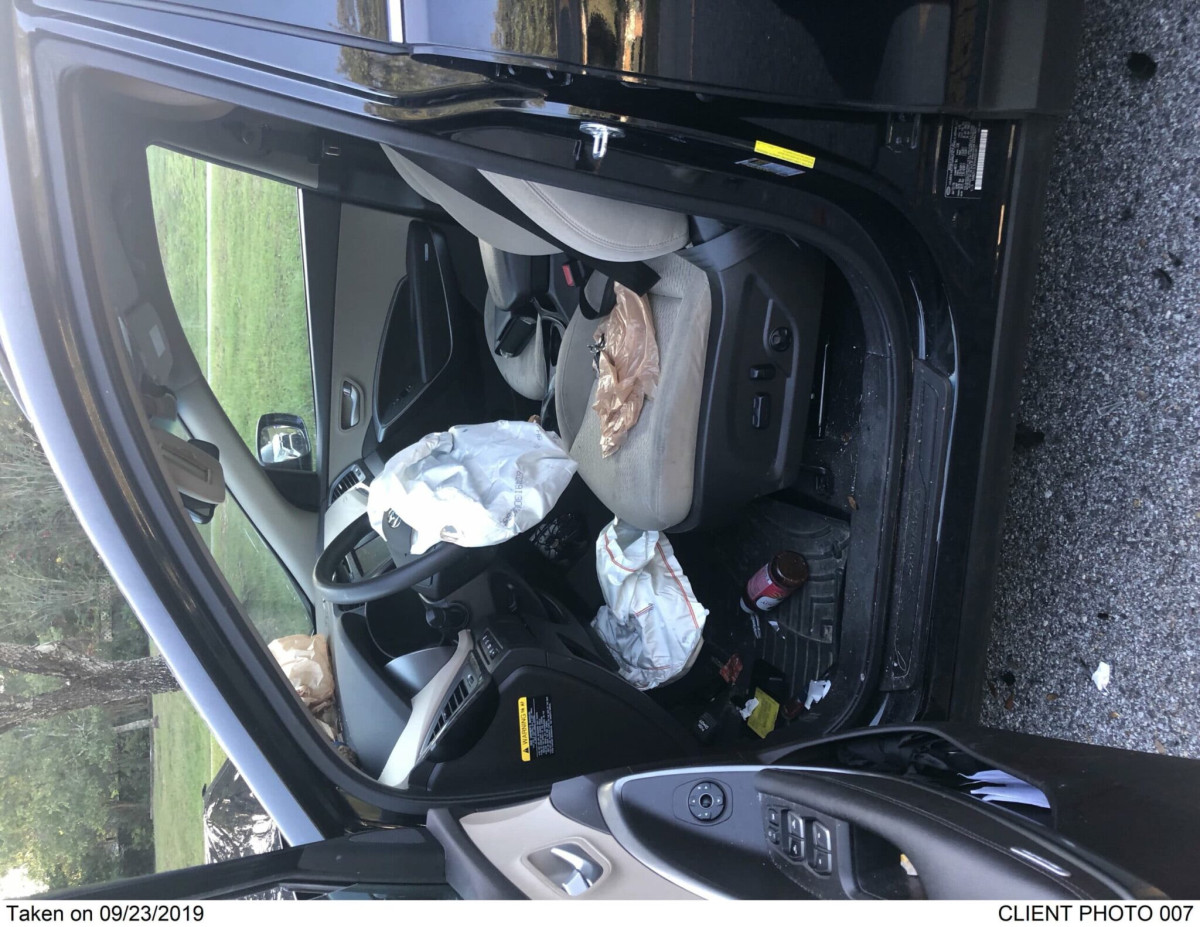 Driver's side view of a car with airbags deployed, plastic bags on the seat, and debris on the floor, suggesting recent impact or accident.