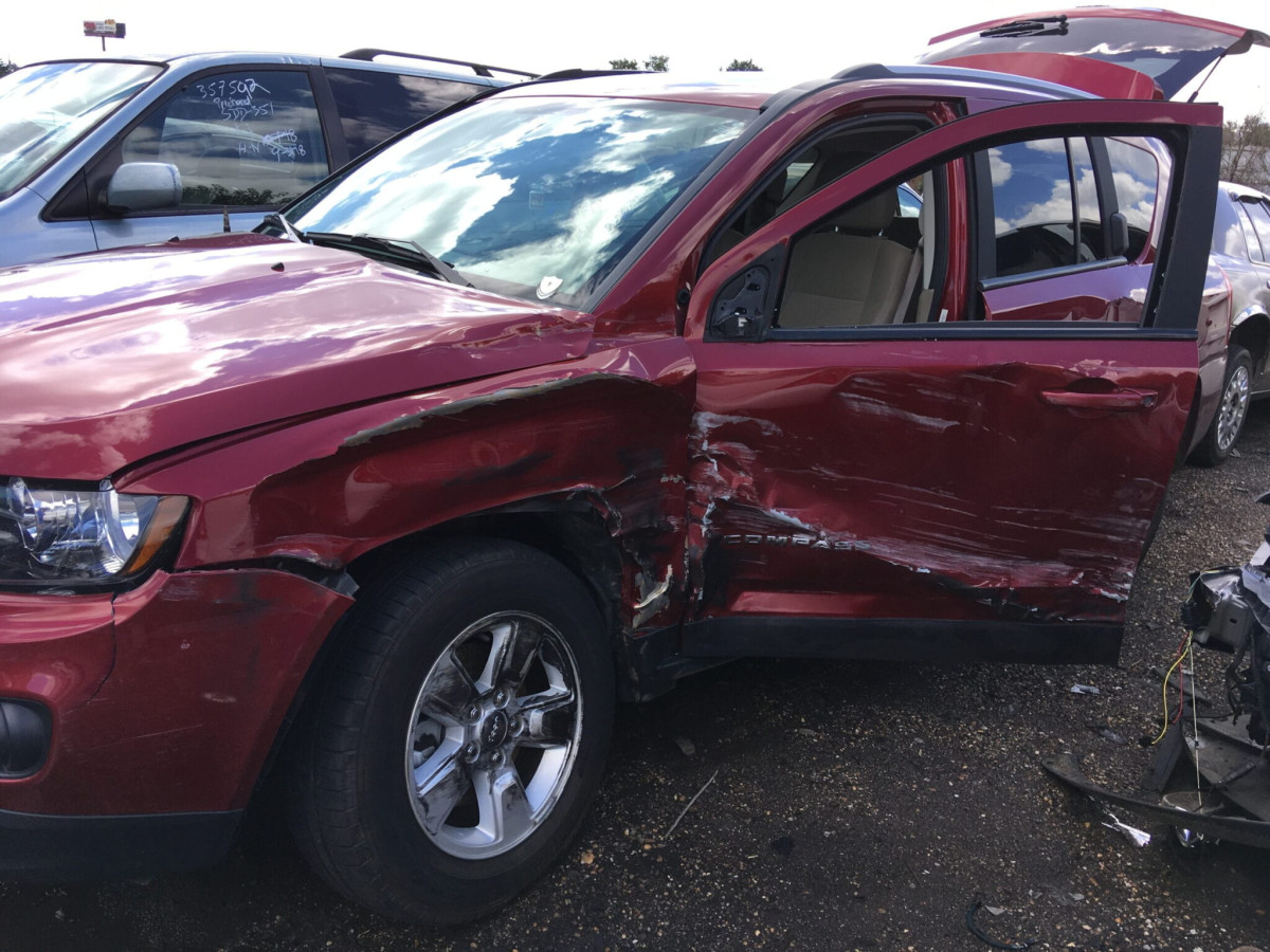 A red SUV with significant damage to the driver's side door and front fender is parked in a lot with its front door open.