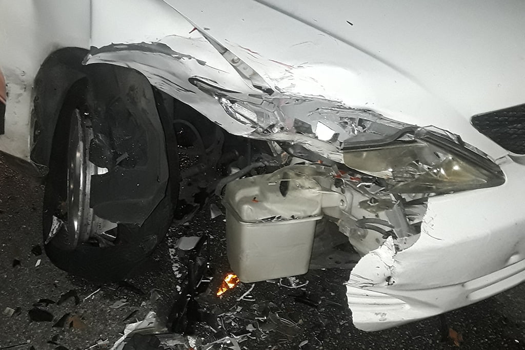 Front left side of a white car damaged from a collision, with a crushed fender, exposed headlight, visible debris, and a bent wheel on the ground.