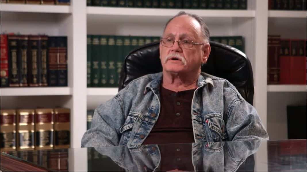 An older man with glasses and a mustache sits in a leather chair, wearing a denim jacket and dark shirt, with bookshelves in the background.