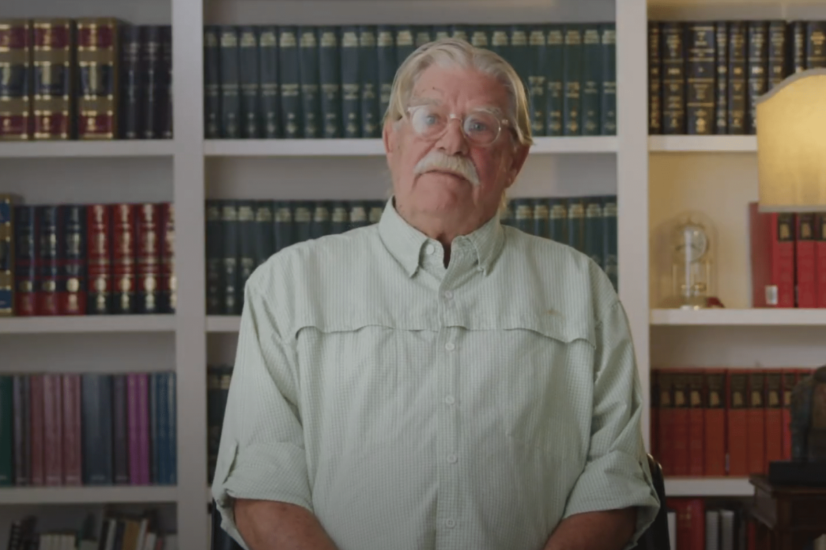 An older man with white hair and glasses sits in front of shelves filled with books, wearing a light green shirt.