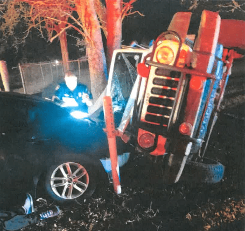 A red Jeep is flipped onto its side against a black car at night, with a police officer standing nearby illuminated by bright lights.