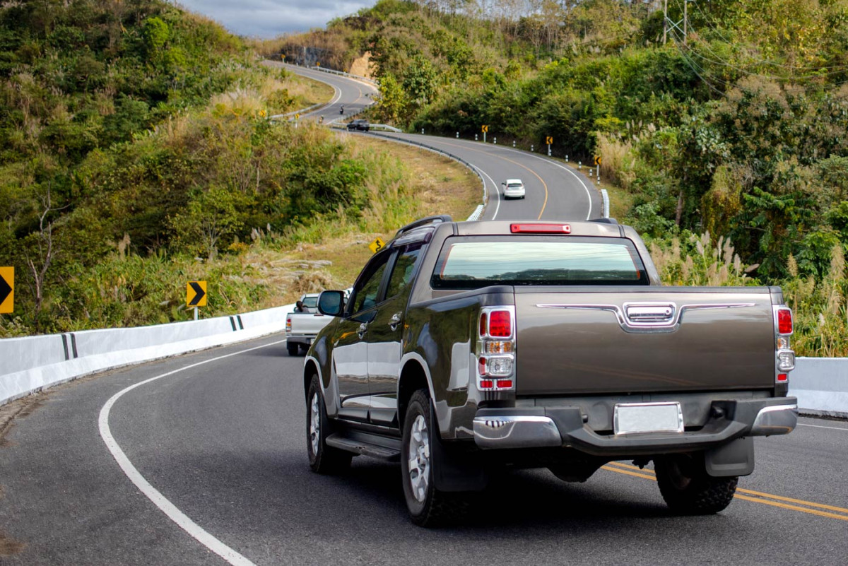 A gray pickup truck drives on a winding road with other cars ahead, surrounded by green hills and vegetation.